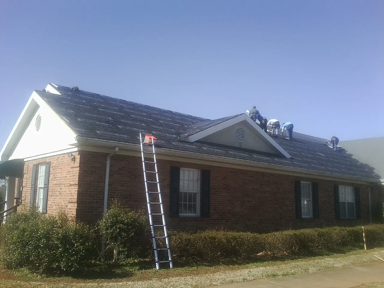Crew tearing shingles off a brick ranch roof with a ladder set — Emergency Roof Repair work by our crew in Eagle Springs
