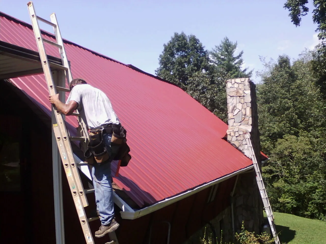 Roofer on a ladder beside a red metal roof and stone chimney — Soffit & Fascia Repair by Roofing Brothers in Eagle Springs